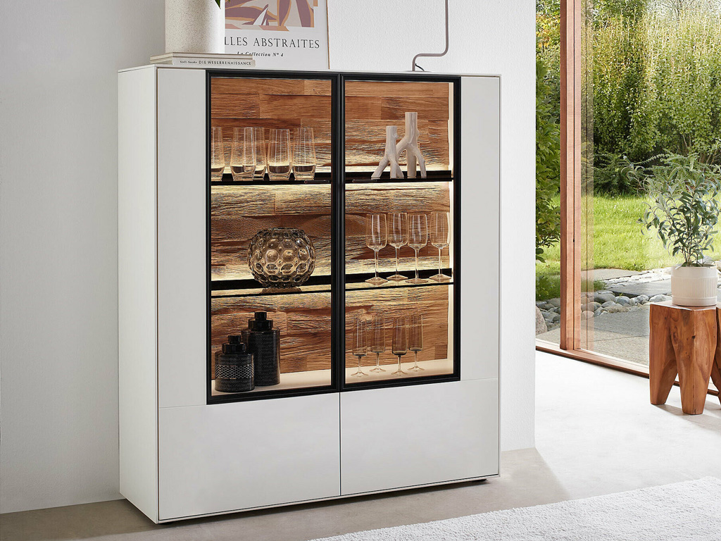 Wöstmann WM 2470 - Highboard | Type 4230 | B: 121,9 cm Weißer Vitrinenschrank mit Glastüren, gefüllt mit Gläsern und Deko, vor einem Fenster mit Gartenblick