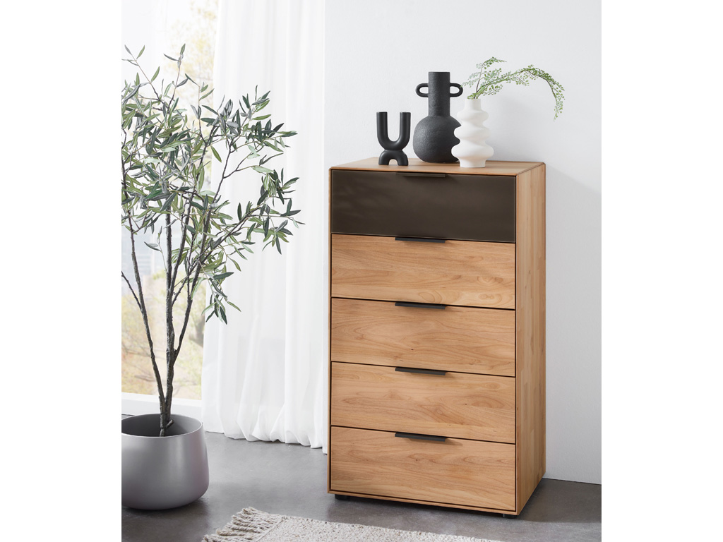 Wöstmann WSM 4400 - Kommode (3652), 5 Schubkästen | Erle massiv | B: 61,5 cm | konfigurierbar Ein moderner Holzschrank mit fünf Schubladen, dekoriert mit Vasen auf der Oberseite, neben einer grünen Pflanze vor einem Fenster.