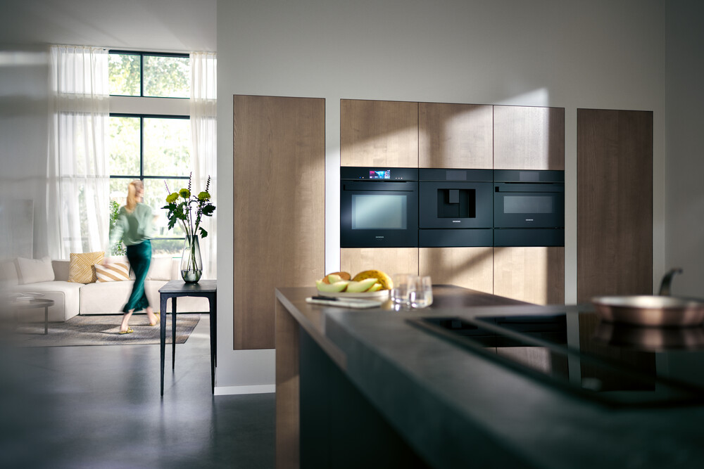 Blick in eine Küche mit hochwertigen Designer-Elektrogeräten mit schwarzer Front und einem Seitenblick in das Wohnzimmer, in dem gerade eine Frau wegläuft.
