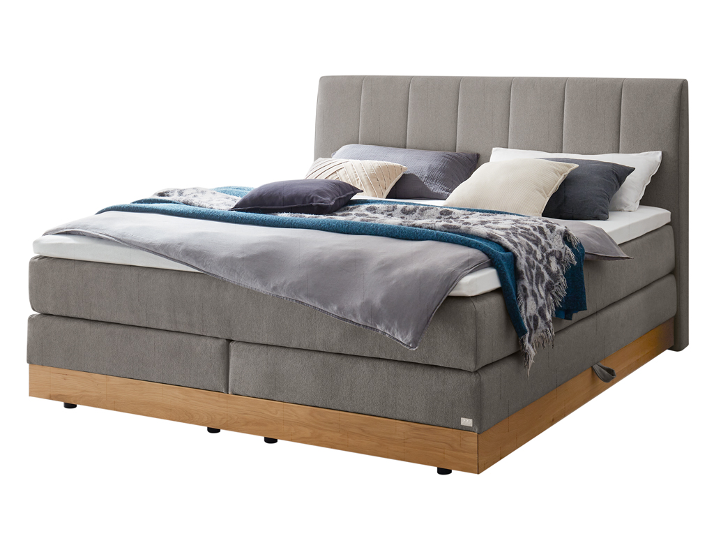 Ein Boxspringbett in mittlerem Grau mit einem gesteppten Kopfteil und einem Holzbettrahmen, freigestellt auf weißem Hintergrund.
