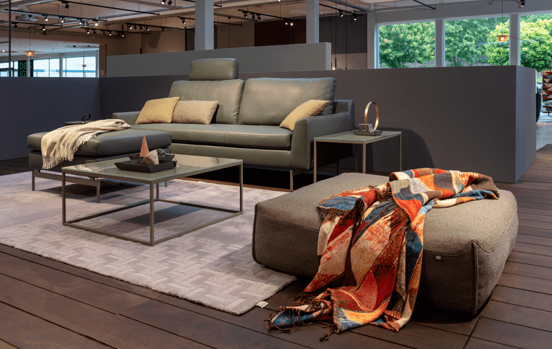 Wohnzimmereinrichtung mit Ledercouch in grau/braun mit passendem Hocker und farbigem Plaid. In der Mitte ein Couchtisch mit Glastischplatte in grau/braun