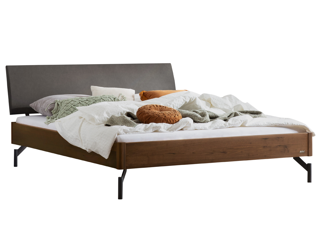 Eine Freisteller-Ansicht des Bettes "Broni" mit einem dunklen Nussbaum-Bettrahmen. Das Bett hat ein anthrazitfarbenes gepolstertes Kopfteil und steht auf schwarzen Metallfüßen. Es ist mit Bettwäsche und Kissen dekoriert.