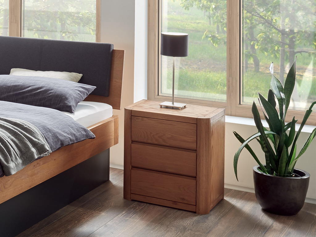 brauner Holznachttisch mit drei Schubladen und abgerundeten Seiten stehend neber einem Holzbett stilvoll dekoriert mit einer Nachttischlampe Koro von Hasena