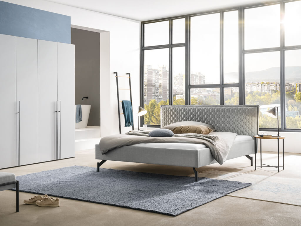 Ein modernes Schlafzimmer mit dem Bett Nardo in einem Loft-Stil, das durch große Fenster und helle Farben eine freundliche Atmosphäre schafft.