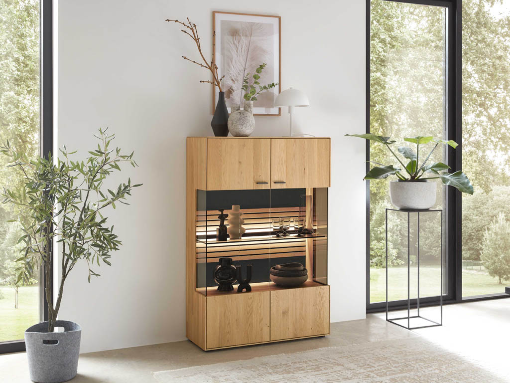 Wöstmann WM 2320 - Highboard  | B: 91,9 cm | Type 4940 Moderner Holzschrank im Wohnzimmer mit Glasregalen, dekoriert mit Vasen und einer Lampe. Umgeben von großen Fenstern und Pflanzen.