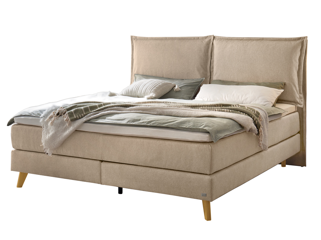 Ein Hasena Boxspringbett Pronto Ricarda mit einem beigefarbenen, sandfarbenen Stoffbezug, das mit Kissen und einer Decke dekoriert ist.