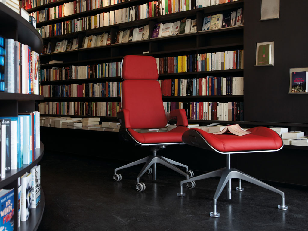 Roter Bürostuhl mit Hocker vor Bücherregal.