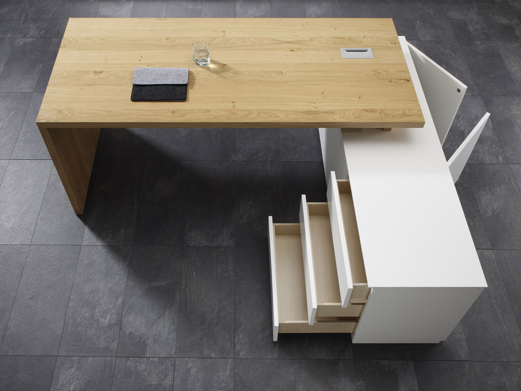Schreibtisch aus Holz mit weißen Schubladen auf dunklem Fliesenboden, darauf ein Laptop und ein Glas Wasser