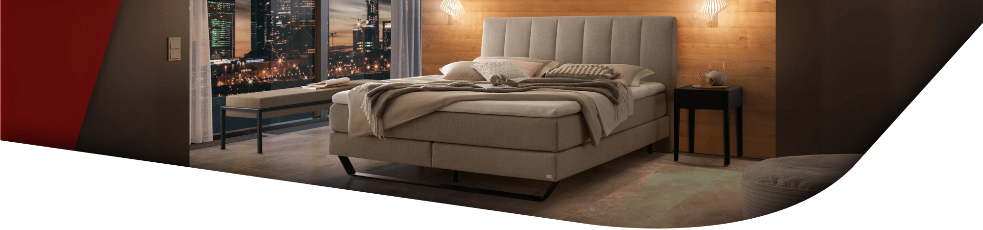 Im Zentrum des Raumes steht ein großes, graues Boxspringbett mit einem gepolsterten Kopfteil. Über den beiden Nachttischen hängen jeweils moderne Pendelleuchten mit holzfarbenen Schirmen.