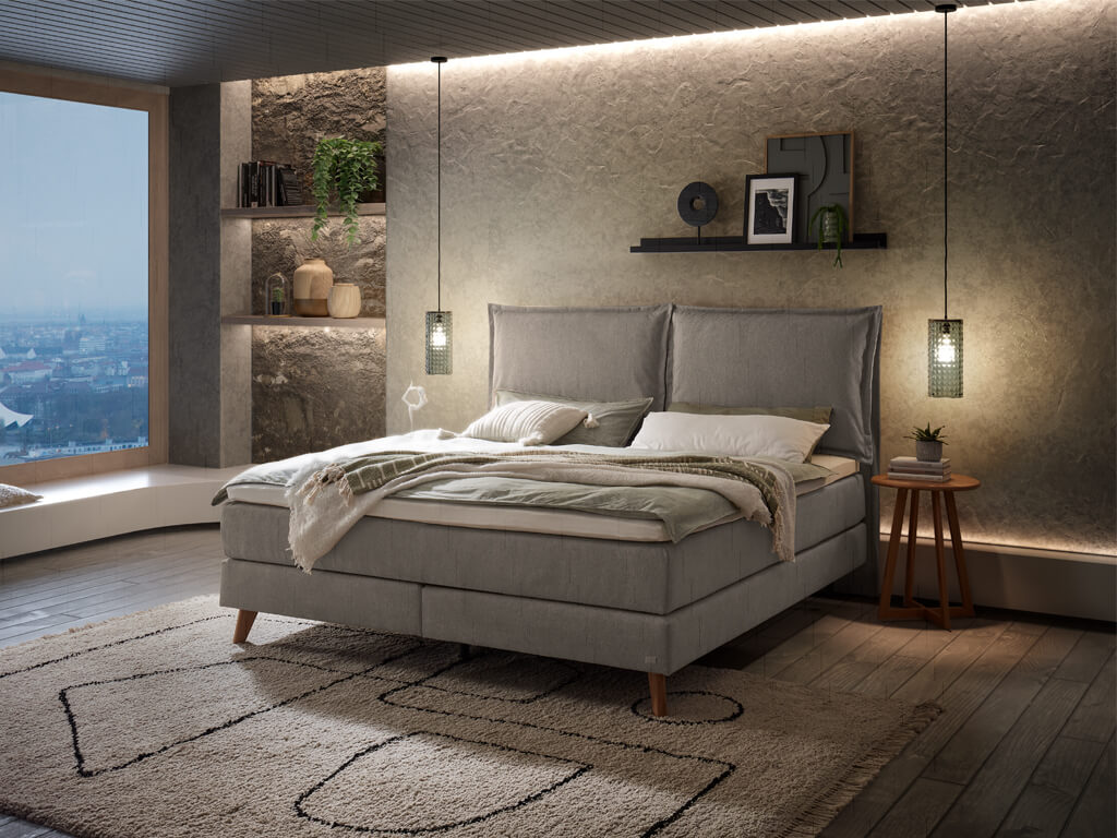 Ein Hasena Boxspringbett Pronto Ricarda Roy in einem modern eingerichteten Schlafzimmer. Das Bett hat einen grauen Stoffbezug und steht auf einem Teppich.
