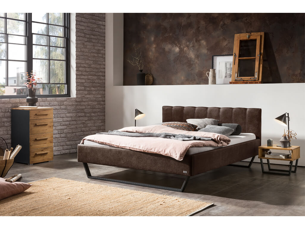 Ein modernes Schlafzimmer mit einem braunen Polsterbett, einem passenden Nachttisch und einer Kommode in einer industriell gestalteten Umgebung