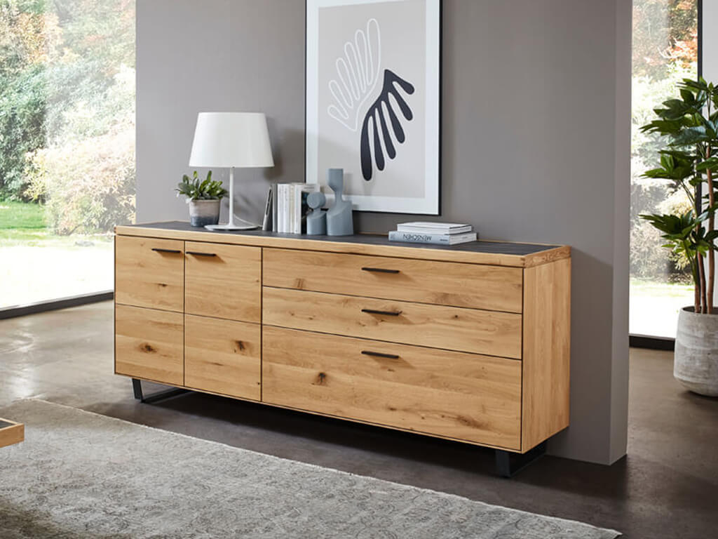 Breites Sideboard aus hellem Holz mit Schubladen und Türen sowie einer dunklen Deckplatte vor einer grauen Wand.