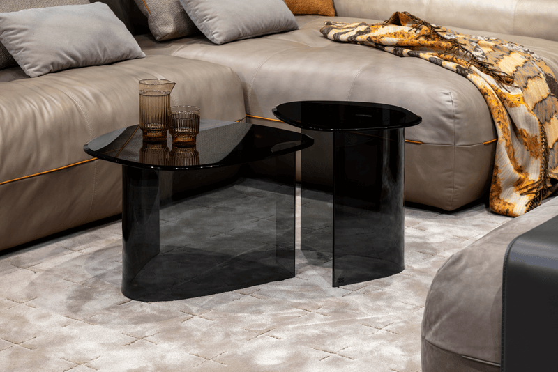 Zwei moderne, schwarze Glastische auf einem hellen Teppich vor einem beigen Ledersofa mit Kissen und einer Decke