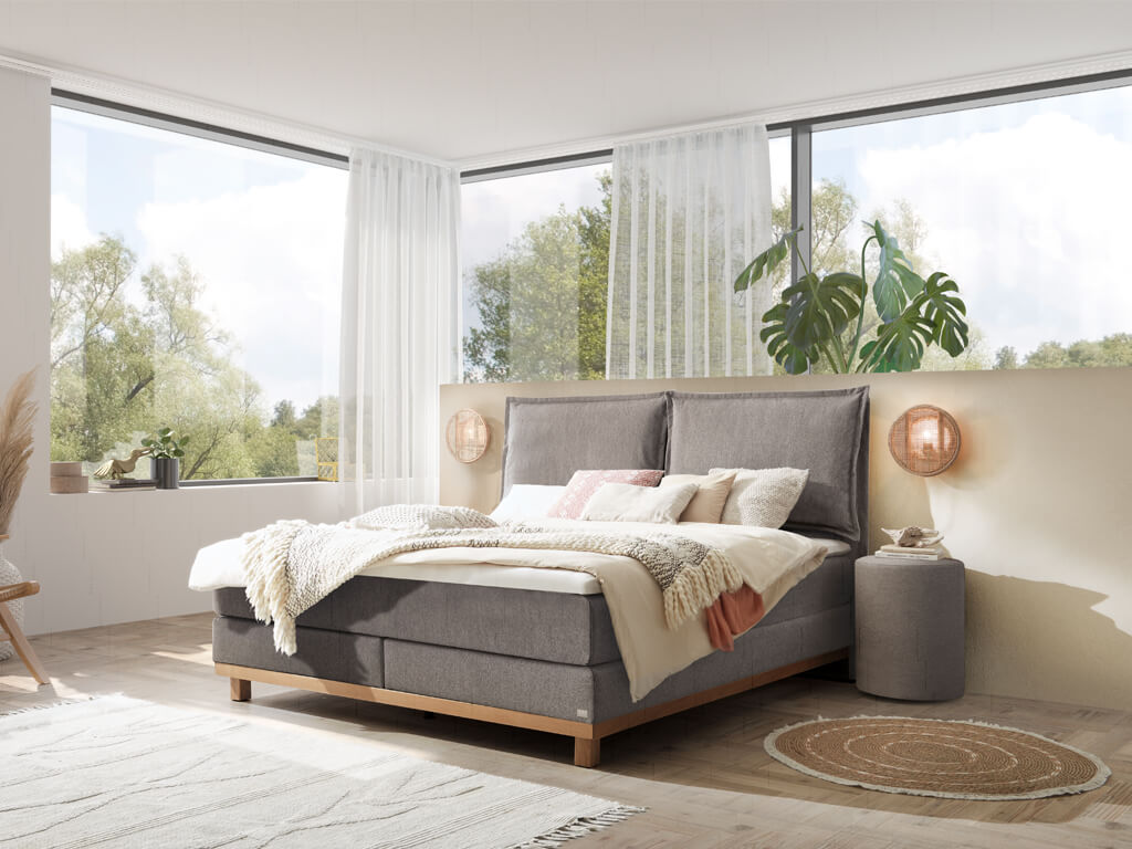 Ein Hasena Boxspringbett Pronto Zamora Dean mit grauem Stoffbezug in einem hellen, von Fenstern durchfluteten Schlafzimmer mit Holzfußboden.