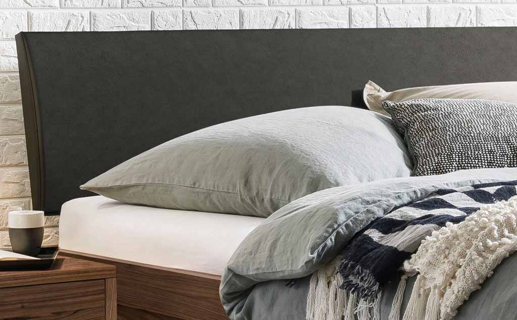 Gemütliches Schlafzimmer mit modernem Bett, grauer Bettwäsche, dekorativen Kissen und Beistelltisch mit Tasse vor einer Ziegelwand