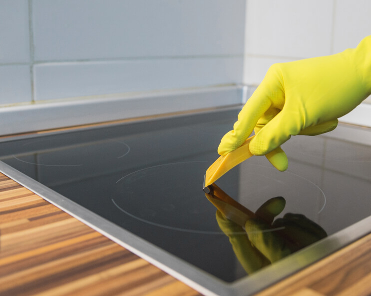 Auf dem Bild sieht man eine Hand ,mit Gummihandschuh, welche miithilfe eines Kochfeldschabers veerkrustungend zu entfernen!