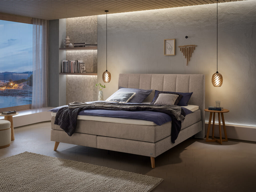 Ein Boxspringbett in beigefarbener Stoffpolsterung und Holzfüßen in einem modern gestalteten Schlafzimmer bei Nacht.