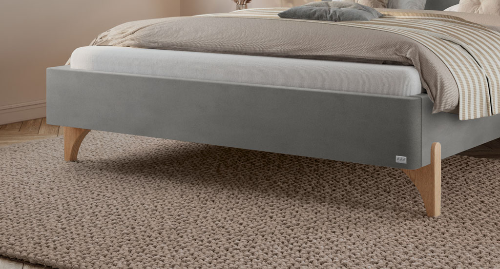 Detailansicht der Eichenholz-Füße an einem grauen Polsterbett auf einem Teppich (KI-generiert)
