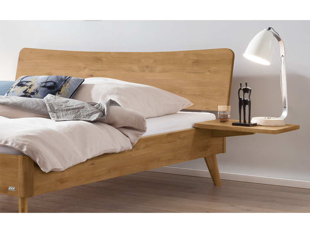 Ein Hasena Bett Caluso aus natur geölter Wildeiche. Das Bett hat ein geschwungenes Kopfteil und schräg ausgestellte Beine. An der Seite ist ein schwebender Nachttisch mit einer Lampe und einer kleinen Skulptur zu sehen.