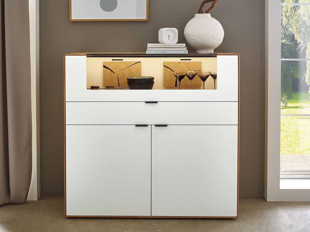 Wöstmann WM 2410 - Highboard | Type 3122 | Lack und Wildeiche massiv | Hirnholzakzent Elegante weiße Kommode mit Glasvitrine, dekorativem Zubehör und stilvoller Beleuchtung in einem Wohnzimmer