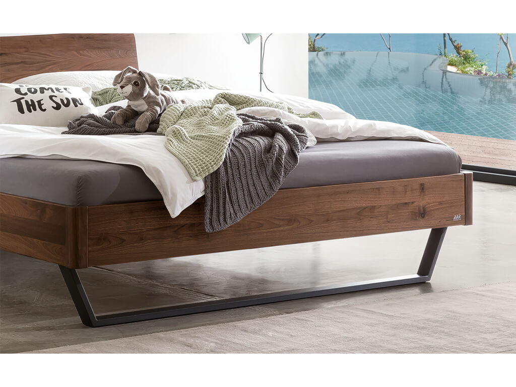 Nahaufnahme der Füße und des Bettrahmens vom Hasena Bett Tresali aus Nussbaum, das vor einem Fenster mit Pool-Blick steht.