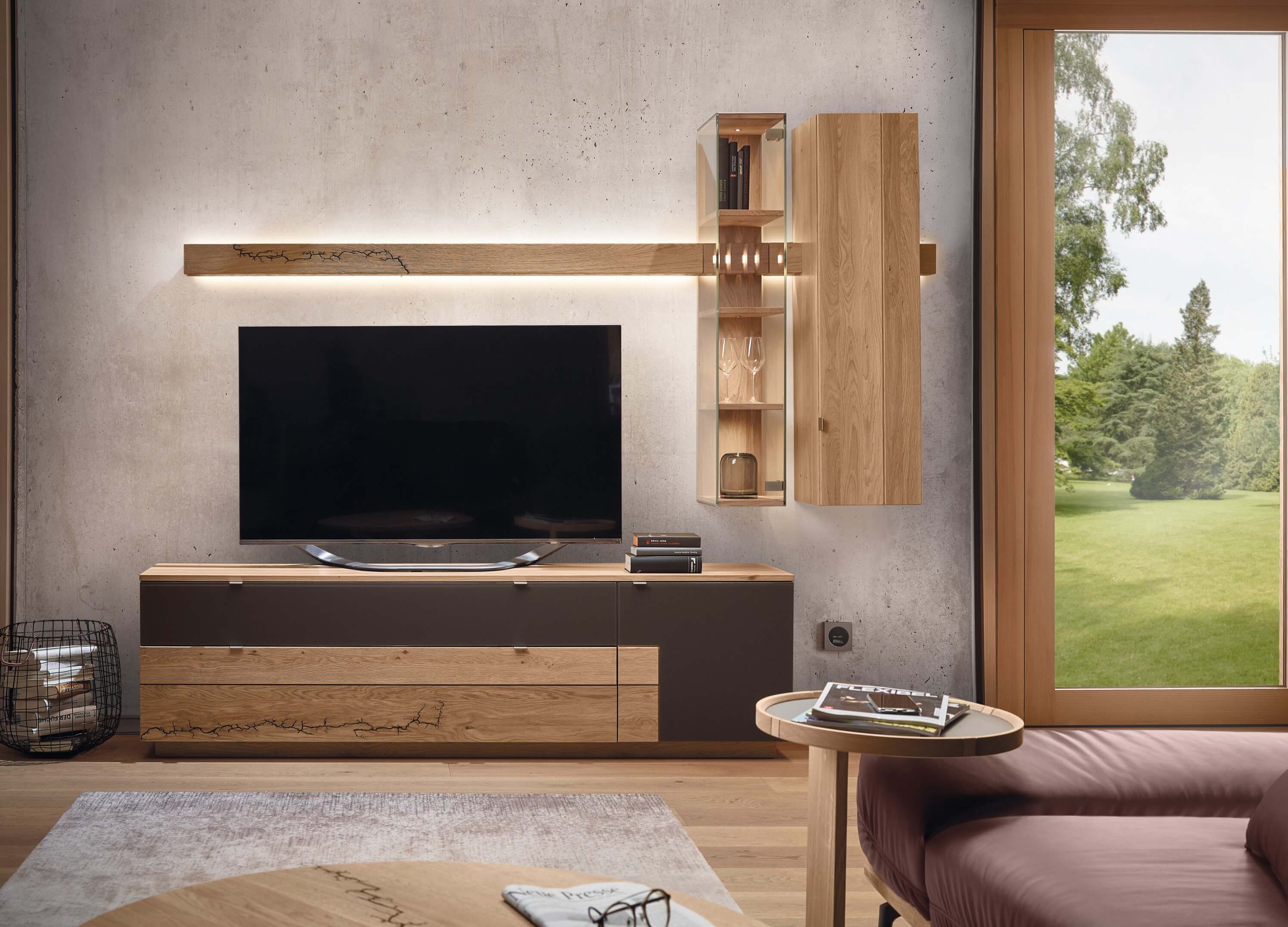 Wimmer Signatura | Wohnwand | 6001140/601141 Ein eleganter Wohnbereich mit einem modernen TV-Schrank aus Holz, akzentuiert durch schicke Regale und eine große Glastür mit Blick ins Grüne.