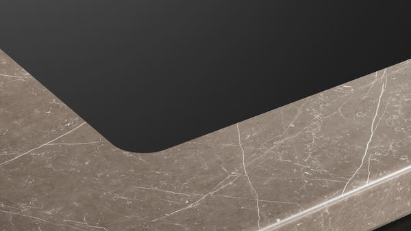 Detailaufnahme der abgerundeten Ecke des schwarzen Induktionskochfelds, das bündig in eine Küchenarbeitsplatte aus braun-grauem Naturstein oder Marmor mit weißen Äderungen eingelassen ist.