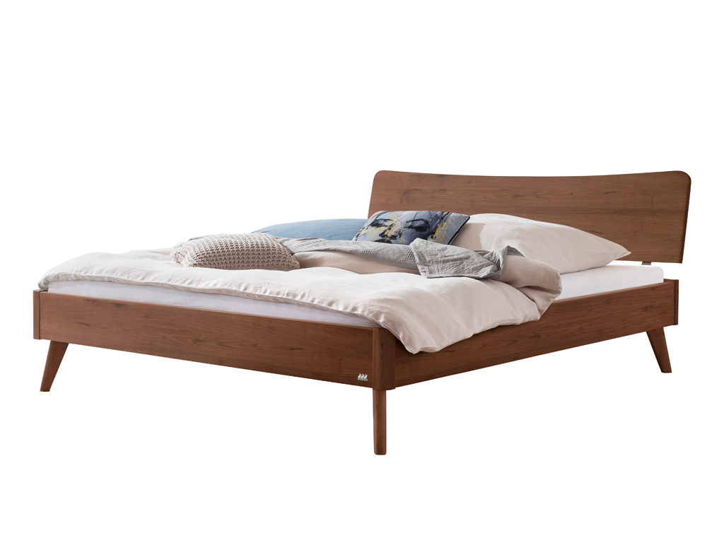 Ein freigestelltes Hasena Bett Caluso aus Nussbaum. Das Bett hat ein geschwungenes Kopfteil, schräg ausgestellte Beine und ist mit Kissen und einer Decke dekoriert.