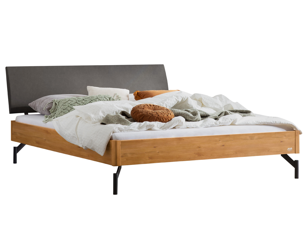 Eine Freisteller-Ansicht des Bettes "Broni" mit einem Bettrahmen aus natur geölter Wildeiche. Es hat ein anthrazitfarbenes gepolstertes Kopfteil und schwarze Metallfüße. Das Bett ist mit Bettwäsche und Kissen dekoriert.