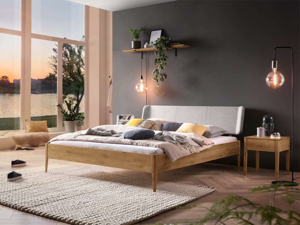 Hasena -  Lounge Bett Pienza | Polsterkopfteil Boga | Holzfuß Pile | versch. Größen & Farben konfigurierbar Schlafzimmer in der Dämmerung mit großer Fensterfront und hellem Holzbett mit hellgrauem Kopfteil