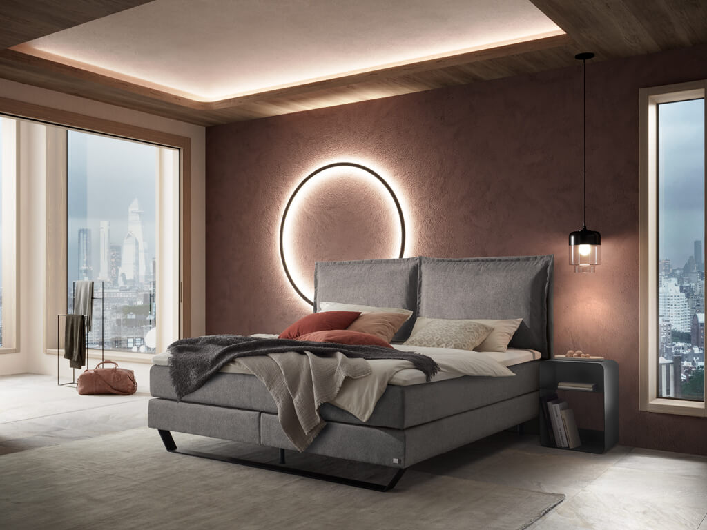 Hasena - Boxspring PRONTO Celina | inkl. Matratze und Topper | versch. Größen & Farben konfigurierbar Ein modernes Schlafzimmer mit einem grauen Hasena Boxspringbett "Pronto Celina" in einem Raum mit rosafarbener Wand und einem großen, beleuchteten Kreis als Wanddeko, aufgenommen aus einem anderen Blickwinkel.
