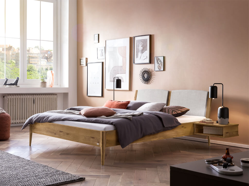 Ein Schlafzimmer mit einem Hasena Pistoia Bett aus natur geölter Wildeiche und gepolstertem, hellgrauen Kopfteil. Es ist ein schwebender Nachttisch integriert und eine moderne Beleuchtung vorhanden.