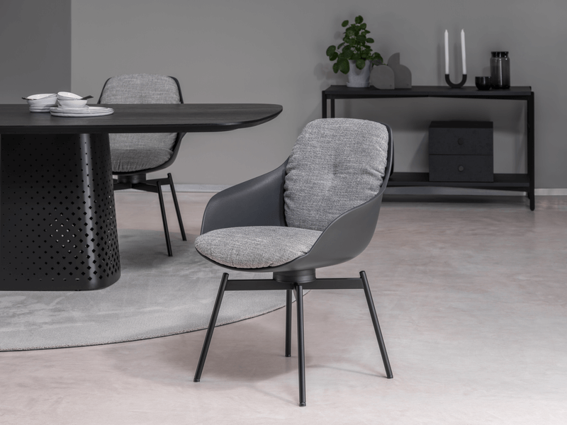 Ein grauer, moderner Esszimmerstuhl steht neben einem schwarzen Esstisch mit dekorativen Löchern. Im Hintergrund befindet sich ein schwarzes Sideboard mit Pflanzen und Kerzen.