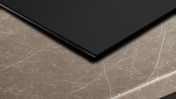 Detailaufnahme der schmalen Kante eines schwarzen Induktionskochfelds, das nahtlos in eine braun-marmorierte Arbeitsplatte eingelassen ist.