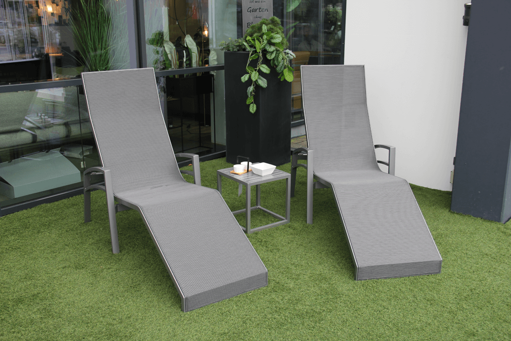 Zwei graue Gartenliegen mit Beistelltisch auf einer Terrasse mit Kunstrasen, Pflanzen im Hintergrund